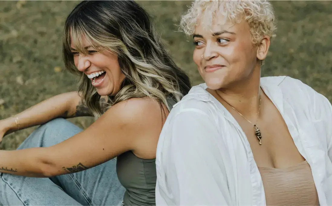 Two women sitting back-to-back, one with wavy brown hair laughing, and the other with short curly blonde hair smiling. They are outdoors on grass, wearing casual attire.