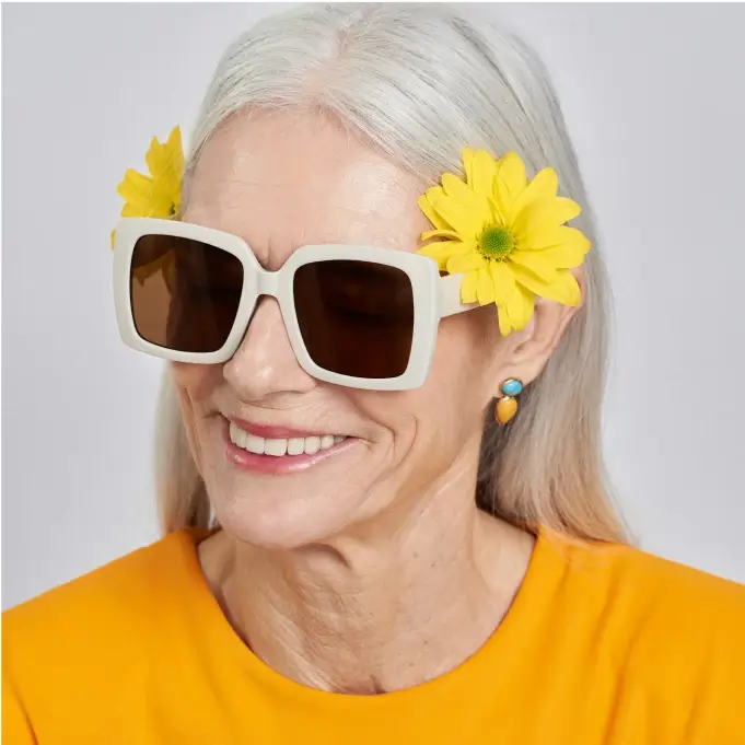 A woman with long white hair is wearing large white sunglasses and has yellow flowers tucked behind her ears. She is smiling and dressed in a bright yellow top.