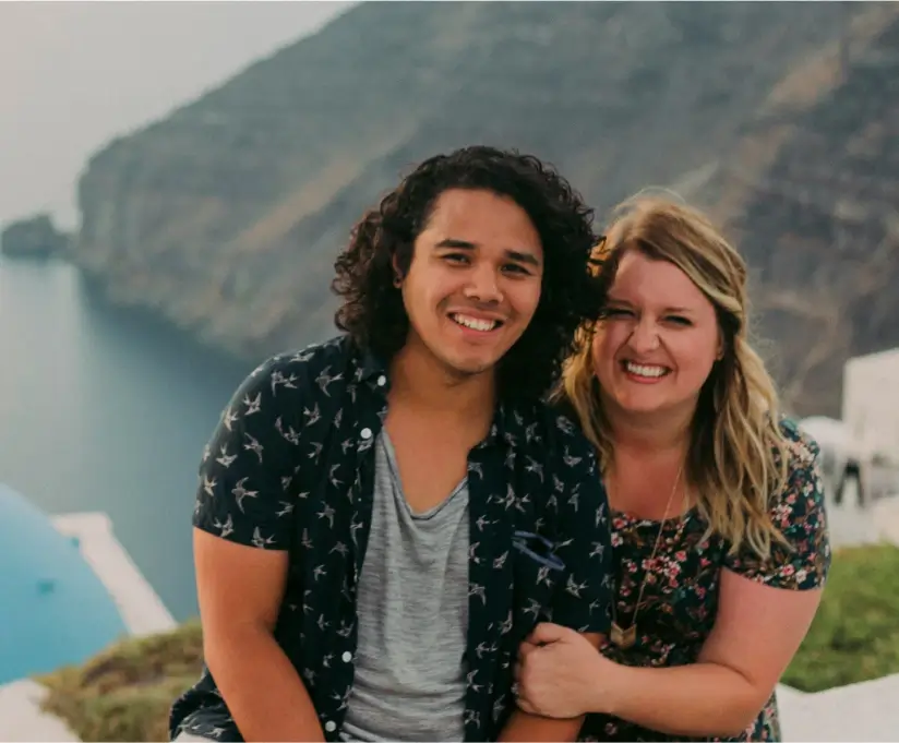 Two people smiling and posing together near a cliffside overlooking the sea with misty mountains in the background.
