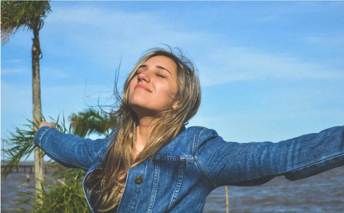 A woman with long hair, wearing a blue denim jacket, stands with her arms outstretched and eyes closed under a blue sky with palm trees in the background.