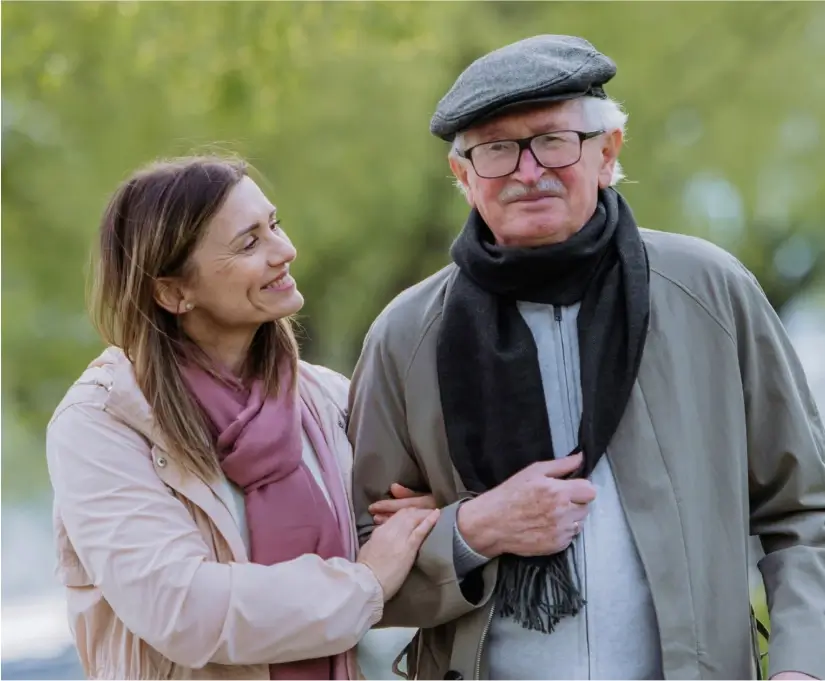 An elderly man wearing a hat and scarf is walking outside with a younger woman who is holding his arm and smiling at him.